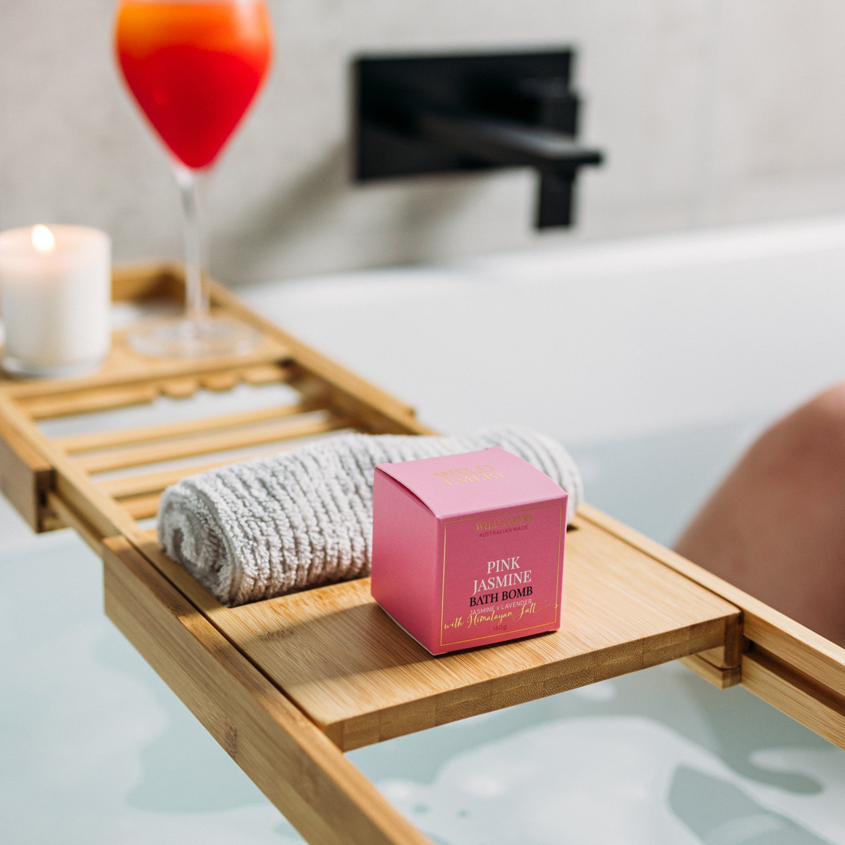 Bath caddy with a glass of red wine, candle, and bath bomb in a bathtub.
