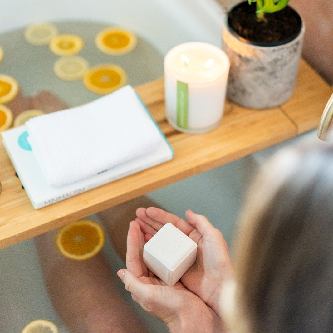 Person holding a bath bomb over a bathtub with floating citrus slices, a candle, and a plant.